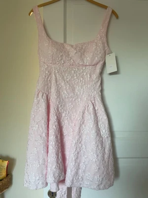 Ljusrosa blommig klänning med spets - Supersöt ljusrosa klänning med blommigt spetsmönster och breda axelband. Klänningen har en markerad midja och ett band som kan knytas till en rosett i ryggen. Perfekt för sommarens alla tillfällen! Det står ingen storlek men passar nog som ungefär en S/M.