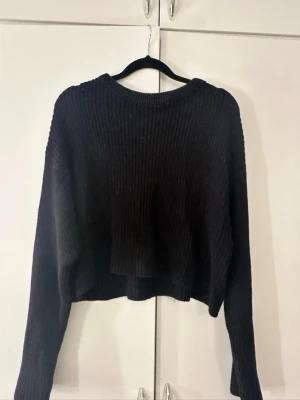 Svart croppad stickad tröja - Säljer en svart croppad stickad tröja med bred ribbstickning och långa ärmar. Tröjan har en avslappnad passform och rund halsringning, perfekt för en trendig och bekväm look.