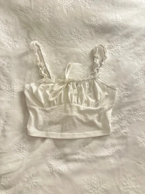 Vit croppad linnetopp med volang - Söt vit croppad linnetopp med volangdetaljer och rynkade axelband. Toppen har en liten rosett framtill och är perfekt för varma dagar. Tillverkad i mjukt bomullstyg för en bekväm känsla.