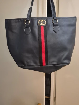 Svart shoppingväska med röd rand - Stilren svart shoppingväska i konstläder med dubbla handtag. Väskan har en dekorativ röd och svart rand framtill samt guldfärgade detaljer och ett emblem med två ringar. Perfekt för dig som vill ha en rymlig och trendig väska till vardagen.