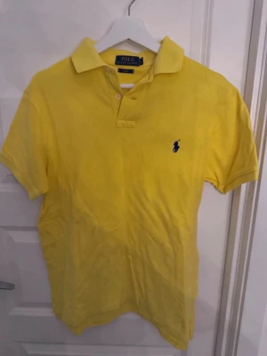 Gul pikétröja från Polo Ralph Lauren - Klassisk gul pikétröja från Polo Ralph Lauren med blå logga broderad på bröstet. Tröjan har korta ärmar, krage och knappar vid halsen. Tillverkad i mjuk bomull som känns skön mot huden. Perfekt för en avslappnad och stilren look.