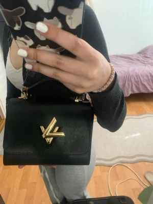 Svart Louis Vuitton handväska - Snygg svart handväska från Louis Vuitton med guldiga metalldetaljer och LV-logga framtill. Väskan har en stilren, fyrkantig form och är tillverkad i strukturerat läder. Insidan har röd detalj och väskan har både handtag och axelrem. Den är äkta men har haft den en bra tag så vill sälja den om du har en fråga dm mig💕dessutom pris kan diskuteras. Kvittot saknas eftersom jag har haft den i ett bra tag nu.
