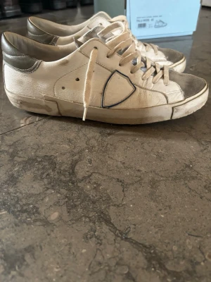 Vita Philippe Model sneakers - Snygga vita sneakers från Philippe Model med grå detaljer och klassisk låg siluett. Skorna har en ikonisk sköldlogga på sidan, vita snören och kontrasterande grå hälkappa i läder. Ovandelen är i textil och mocka för en trendig look.