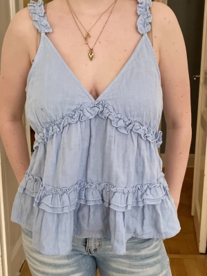 Ljusblå volangtop med v-ringning - Supersöt ljusblå linnetopp med volanger och v-ringning. Axelbanden har volangdetaljer och toppen är ärmlös med en luftig passform. Perfekt för varma dagar och ger en romantisk vibe till din outfit.