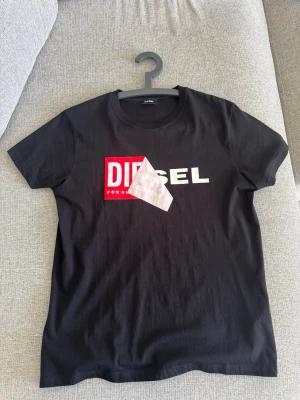 Svart t-shirt från Diesel - Snygg svart t-shirt från Diesel med klassisk logga i vitt och rött framtill. T-shirten har rund halsringning och korta ärmar. Tillverkad i mjuk bomull som känns skön mot huden. Perfekt för en avslappnad och trendig look.