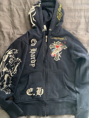  Ed Hardy hoodie med tryck - Snygg mörkblå hoodie från Ed Hardy med dragkedja, stora vita tryck på ärm och bröst samt färgglad korsdetalj framtill. Huva med snörning och Ed Hardy-logga. Perfekt för dig som gillar streetwear och unika detaljer. (Tröjan har nån fläck av nagellack som syns på sista bilden, vet ej om de går bort har inte testat)💕  Nyköpt för 1300kr ❤️ 