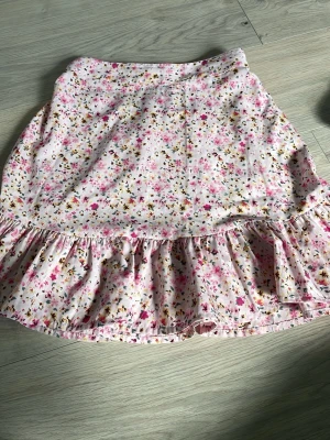 Blommig volangkjol från Vero Moda - Supersöt kort kjol från Vero Moda med blommigt mönster i rosa, gult och grönt på ljus botten. Kjolen har volangkant nedtill och resår i midjan för skön passform. Perfekt för dig som gillar en feminin och lekfull stil.