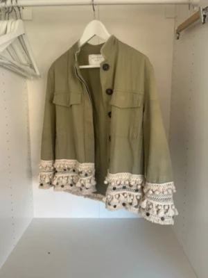 Grön jacka med virkade detaljer - Snygg grön jacka med virkade och broderade detaljer samt tofsar i beige längs ärmslut och nederkant. Jackan har knappar framtill och två bröstfickor. Perfekt för dig som vill sticka ut med en unik stil. Är i storlek M men sitter mer som S