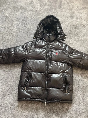 Svart pufferjacka från Polo Ralph Lauren - Snygg svart pufferjacka från Polo Ralph Lauren med blank finish och röd logga på bröstet. Jackan har huva, två dragkedjefickor och breda kanaler för extra värme. Perfekt för kalla vinterdagar och riktigt stilren look.