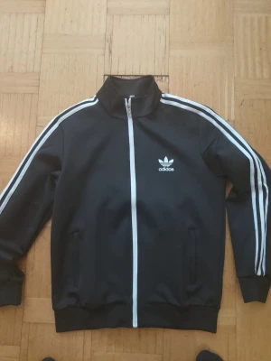 Svart Adidas track jacket - Svart track jacket från Adidas med klassiska vita ränder längs ärmarna och vit dragkedja. Jackan har hög krage, muddar vid ärmslut och nederkant samt Adidas logga på bröstet. Perfekt för sport eller chill. Materialet är polyester. Storleken säger XXL men den passar även XL