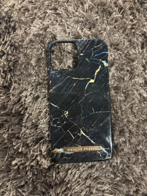 Marmorerat mobilskal Ideal of Sweden iPhone 12 - Snyggt mobilskal från Ideal of Sweden med svart marmormönster och inslag av guld och blått. Skalets yta är slät och har en exklusiv känsla. Passar perfekt för dig som vill ha ett stilrent och trendigt skydd till din mobil. Pris kan diskuteras 