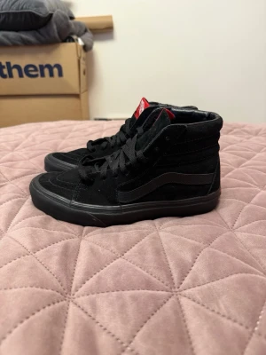 Svarta Vans Sk8-Hi sneakers - Svarta Vans Sk8-Hi sneakers med högt skaft, klassisk vågig sidorand och röd logga på plösen. Skorna är gjorda i mocka och canvas med svarta skosnören och gummisula. Perfekta för en streetwear-look.