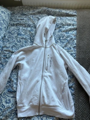 Vit hoodie från Sailracing  - Säljer en vit hoodie från Sailracing med dragkedja framtill och en extra ficka med dragkedja på bröstet. Insidan är mjuk och fleecefodrad för extra komfort. Klassisk huva med snörning och ribbade muddar vid ärmslut och nederkant.