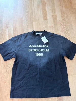 Svart Acne Studios t-shirt med tryck - Svart t-shirt från Acne Studios med vit text 'Acne Studios STOCKHOLM 1996' på bröstet. Klassisk passform med rund hals och korta ärmar. Perfekt för en stilren och trendig look.