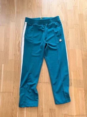 Moncler X palm angels mjukisbyxor med vit rand - Snygga turkosa mjukisbyxor från Moncler med vita ränder längs sidorna och elastisk midja. Byxorna har raka ben och två fickor framtill. Perfekta för en chill och sportig look. Storlek L baggy fit