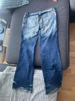 Blå bootcut jeans med broderi - Säljer ett par blå bootcut jeans från Evisu med ljusa slitningar och broderade detaljer på ena benet. Jeansen har klassisk femficksdesign och synliga slitningar vid benslut. Perfekta för dig som gillar en avslappnad och trendig stil.