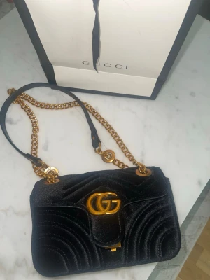 Svart sammetsväska från Gucci - Snygg svart handväska från Gucci i mjuk sammet med guldfärgade metalldetaljer och kedjerem. Väskan har GG-logga framtill och ett innerfack med dragkedja. Perfekt för dig som vill ha en lyxig och trendig accessoar.