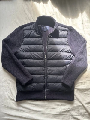 Mörkblå Cardigan Leon Marcél - Stilren mörkblå cardiganjacka från Leon Marcel med vadderad framsida i nylon och stickade ärmar och rygg i bomull, ull och kashmir. Jackan har dragkedja framtill och ribbade muddar. Perfekt för lager-på-lager och en clean look.
