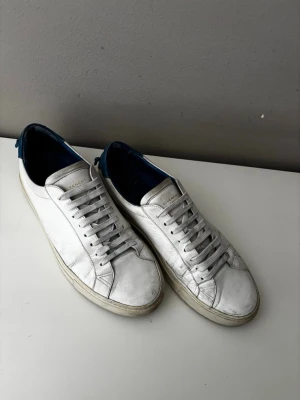 Vita Givenchy sneakers med blå detalj - Snygga vita sneakers från Givenchy med blå hälkappa och guldfärgad logga. Skorna har klassisk låg profil, rund tå och är tillverkade i läder. Perfekta för dig som gillar stilrena och exklusiva sneakers med en twist.