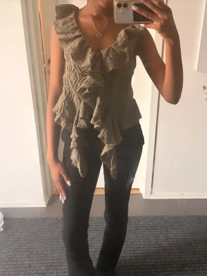 Beige volangblus med omlottkänsla - Supersnygg beige blus med volangdetaljer och omlottkänsla. Ärmlös modell med v-ringning och strukturerat tyg som ger en cool look. Perfekt att styla med jeans eller kjol för en trendig outfit.
