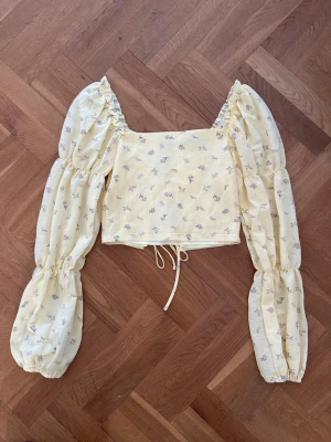 Gul blommig blus med puffärmar - Supersöt gul blus med små blommor och stora puffärmar. Toppen har snörning både fram och bak samt en croppad passform. Perfekt för dig som gillar romantisk och trendig stil. Materialet känns lätt och luftigt.