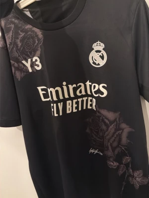 Real Madrid x Y-3 svart matchtröja - Svart Real Madrid matchtröja i samarbete med Y-3, med vita tryck och detaljer som klubbmärke, sponsorlogga och stora rosor på axel och nedre del. Tillverkad i funktionsmaterial, perfekt för fotboll eller träning.