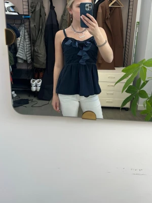 Mörkblå volangtop med rosett - Supersöt mörkblå topp med volanger och rosett framtill. Axelbandsmodell med luftig passform och lätt utsvängd nederdel. Perfekt för dig som vill ha en feminin och trendig look.