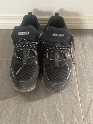 Svarta Lacoste sneakers med meshdetaljer - Snygga svarta sneakers från Lacoste med coola meshpaneler och reflekterande detaljer. Skorna har svartvita snören och en robust sula som ger en sportig vibe. Perfekta för dig som gillar streetstyle och vill ha något bekvämt och trendigt på fötterna.