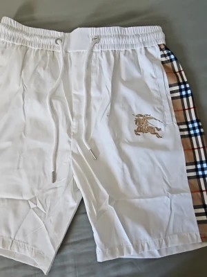Vita shorts med snörning och rutiga sidor - Säljer ett par vita shorts från Burberry med snörning i midjan och klassiska rutiga paneler på sidorna. Shortsens material känns mjukt och lätt, perfekt för varma dagar. Burberry-loggan är broderad på framsidan.