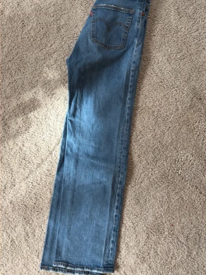 Levi's raka blå jeans ankel storlek 27 - Klassiska raka jeans från Levi's i ljusblå denim. Jeansen har fem fickor, synliga sömmar och den ikoniska röda Levi's-taggen på bakfickan. Perfekta för en avslappnad och tidlös stil.