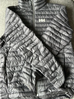Grå pufferjacka från Helly Hansen - Snygg grå pufferjacka från Helly Hansen med hög krage och dragkedja framtill. Jackan har quiltad design, två sidofickor med dragkedja och HH-logga på bröstet. Perfekt för kalla dagar och riktigt lätt att bära. Materialet är syntetiskt och håller värmen bra.
