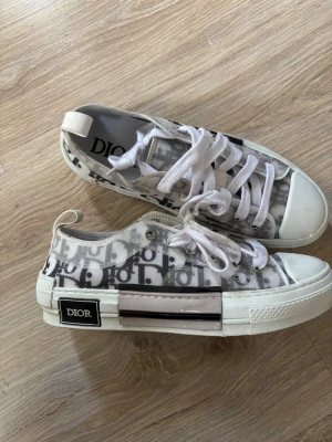 Dior B23 vita  - Snygga Dior B23 sneakers i vitt med genomskinliga paneler och klassiskt Dior-monogram. Skorna har vita snören, gummisula med Dior-logga och detaljer i svart och silver. Utan box, Perfekta för dig som vill sticka ut med exklusiva sneakers. Passar39/40
