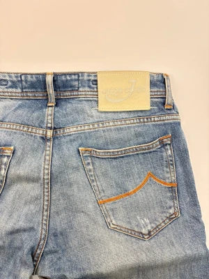 Blå jeans från Jacob Cohen - Snygga blå jeans från Jacob Cohen med klassisk femficksdesign och orange kontrastsömmar på bakfickorna. Jeansen har en rak passform och är tillverkade i mjuk denim med lätt slitna detaljer. Perfekta för en avslappnad och trendig look.
