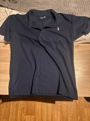 Marinblå pikétröja från Ralph Lauren - Klassisk marinblå pikétröja från Polo Sport Ralph Lauren med korta ärmar och vit broderad logga på bröstet. Tillverkad i 100% bomull och har en snygg krage med knappar. Perfekt till en avslappnad men stilren look.