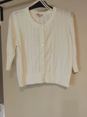 Vit stickad kofta med mönster - Snygg vit stickad kofta från Lindex med diskret diamantmönster och rund halsringning. Koftan har knappar framtill och 3/4-ärmar. Perfekt att slänga över en topp för en clean look. Materialet är mjukt och bekvämt.