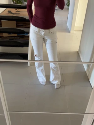 Vita slitna bootcut jeans - Säljer ett par vita bootcut jeans med slitningar och hål på ena benet. Jeansen har klassisk femficksdesign och råa kanter längst ner. Perfekta för dig som gillar en avslappnad och trendig look. Från Gina i S , skriv för fler frågor! 