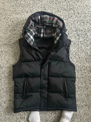 Burberry   - Svart dunväst med huva och rutigt foder i grått, svart och rött. Västen har två dragkedjefickor framtill och stängs med både dragkedja och knappar. Perfekt för lager-på-lager och chill höststil.