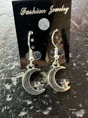 Silvriga örhängen med måne och stjärna - Coola silvriga örhängen i metall med hängande måne och liten stjärna. Snyggt och trendigt motiv som passar perfekt till din stil. Krokar i silverfärgad metall för enkel användning.