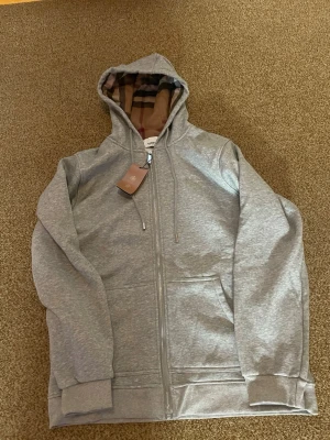 Grå zip hoodie från Burberry - Snygg grå zip hoodie från Burberry med klassiskt rutigt foder i huvan. Hoodien har dragkedja framtill, två fickor och ribbade muddar vid ärmar och nederkant. Perfekt för en chill och stilren look. Materialet känns mjukt och bekvämt.