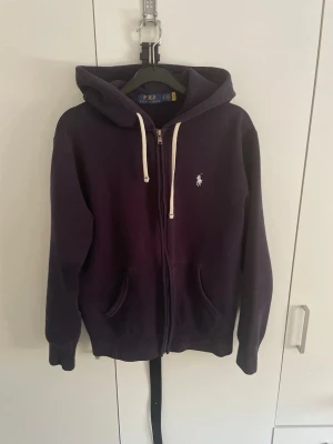 Mörkblå hoodie från Polo Ralph Lauren - Säljer en mörkblå hoodie från Polo Ralph Lauren med dragkedja, vit dragsko och klassisk logga broderad på bröstet. Hoodien har två fickor framtill och är tillverkad i mjukt bomullsmaterial. Perfekt för en chill och stilren look.