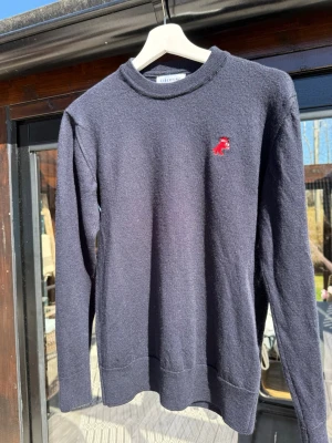 Stickad tröja Libertine Libertine  - Mörkblå stickad tröja från Libertine Libertine i ull med rund halsringning och en liten röd broderad detalj på bröstet. Tröjan har långa ärmar och ribbade muddar vid ärmslut och nederkant. Perfekt för lager-på-lager-stil. Perfekt skick, det är stolen M men passar XS eller S då den har krympt.