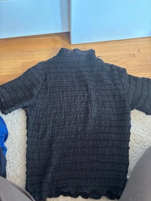 Svart ribbad topp från Stories - Svart ribbad topp från Stories i storlek M. Toppen har korta ärmar, hög hals och ett smockat, stretchigt tyg som ger en cool struktur. Perfekt att styla med jeans eller kjol för en trendig look.