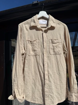 Beige Skjorta från Selected Homme - Stilren beige skjorta från Selected Homme med två bröstfickor och klassisk krage. Skjortan är långärmad och tillverkad i ett mjukt linne och bommulstyg, perfekt för en avslappnad look. Storlek S och perfekt skick. Skriv vid frågor!
