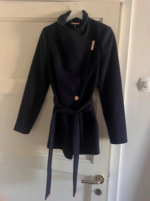Mörkblå Ted Baker-kappa med skärp - Stilren mörkblå kappa med asymmetrisk knäppning och matchande midjeskärp. Kappan har en dekorativ knapp framtill och bred krage som kan bäras både öppen och stängd (dold magnet som gör att den kan vara stängd). Perfekt för dig som gillar minimalistisk design och vill ha en snygg jacka till hösten. Mycket bra skick, nypris ca 4749 kr.