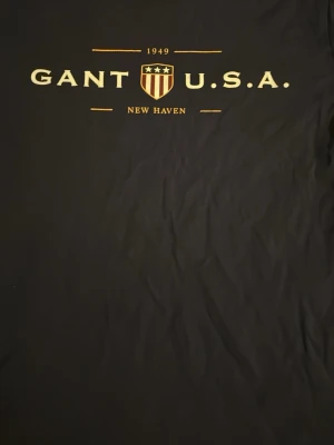 Svart GANT t-shirt med tryck - Snygg svart t-shirt från GANT med klassiskt tryck framtill: 'GANT U.S.A. 1949 New Haven' och sköld. T-shirten har rund hals och är perfekt för en stilren och sportig look.