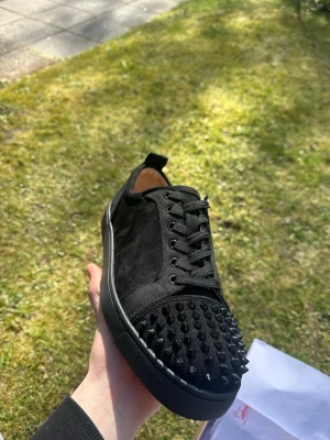 Svarta sneakers med nitar Christian Louboutin - Säljer ett par svarta sneakers från Christian Louboutin med mattsvarta nitar på tån och ovandel i mocka. Skorna har svarta skosnören, svart sula och en klassisk låg silhuett. Perfekta för dig som vill sticka ut med en edgy look.