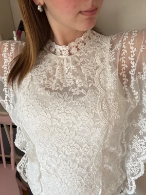 Vit spetsblus med volangdetaljer🫶🏻 - Superfin vit blus i spets med blommigt mönster och volangdetaljer längs ärmarna. Hög hals med söt spetskant och transparenta partier på axlar och ärmar. Perfekt för dig som gillar romantisk och trendig stil. Liten fläck vid halsen (se bild 4), går säkert bort med något bra medel, har bara inte försökt🤍