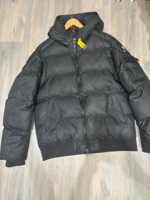 Svart pufferjacka från Parajumpers - Säljer en svart pufferjacka från Parajumpers i storlek L. Jackan har en hög krage, två stora fickor framtill och en ärmficka med logopatch. Materialet är slitstark nylon med fleecefoder och coola orange detaljer. Perfekt för kalla vinterdagar.