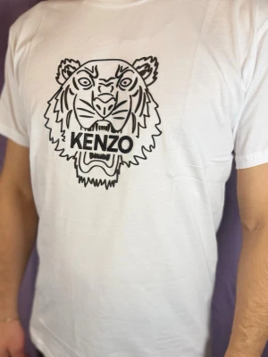 Vit Kenzo t-shirt med tigertryck - Säljer en vit t-shirt från Kenzo med ikoniskt svart tigertryck på bröstet. T-shirten har klassisk passform och korta ärmar, perfekt för dig som vill sticka ut med ett coolt motiv. Materialet är mjuk bomull som känns skönt mot huden.