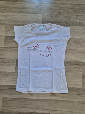 Vit t-shirt med fjärilar och text - Vit t-shirt med korta ärmar och rund halsringning. Framsidan har dekorativa fjärilar och text i glittriga stenar. Materialet är mjuk bomull och passformen är normal. Perfekt för dig som gillar enkel stil med lite extra detaljer.